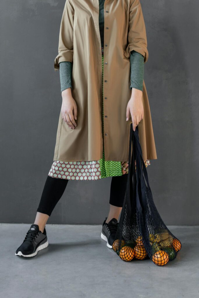 A stylish outfit featuring a trench coat and sneakers with a reusable net bag and fruits.